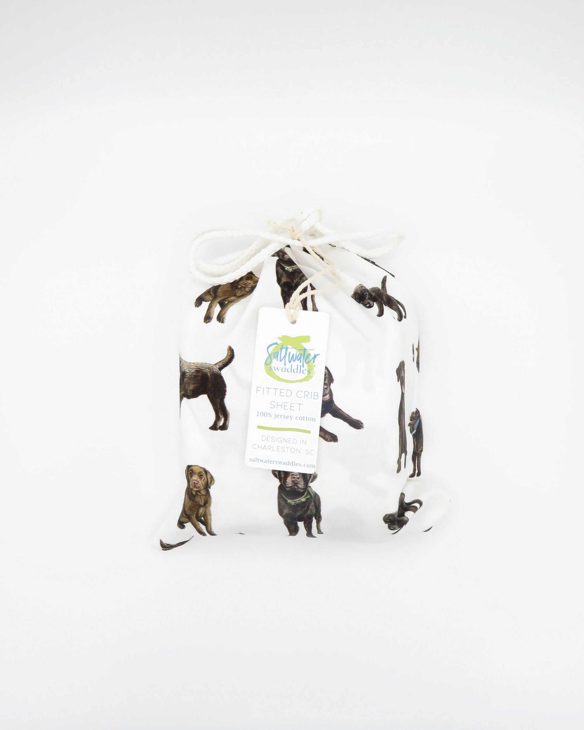 A white crib sheet with a chocolate labrador retriever print, displayed in packaging bag tied with a ribbon on a white background.