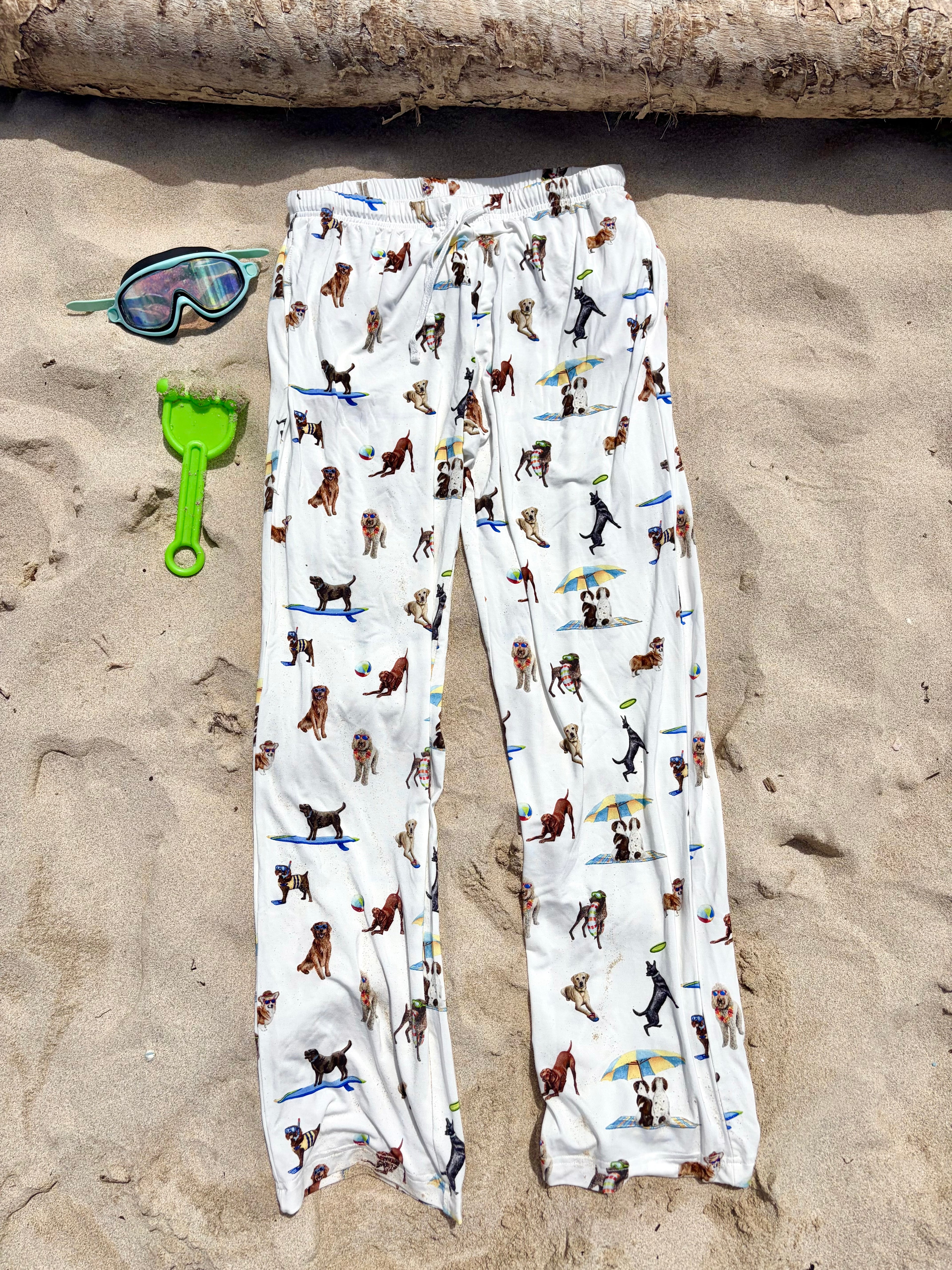 Dog Days of Summer adult pajama pants displayed flat on the beach sand, with kids’ swimming accessories on the side.