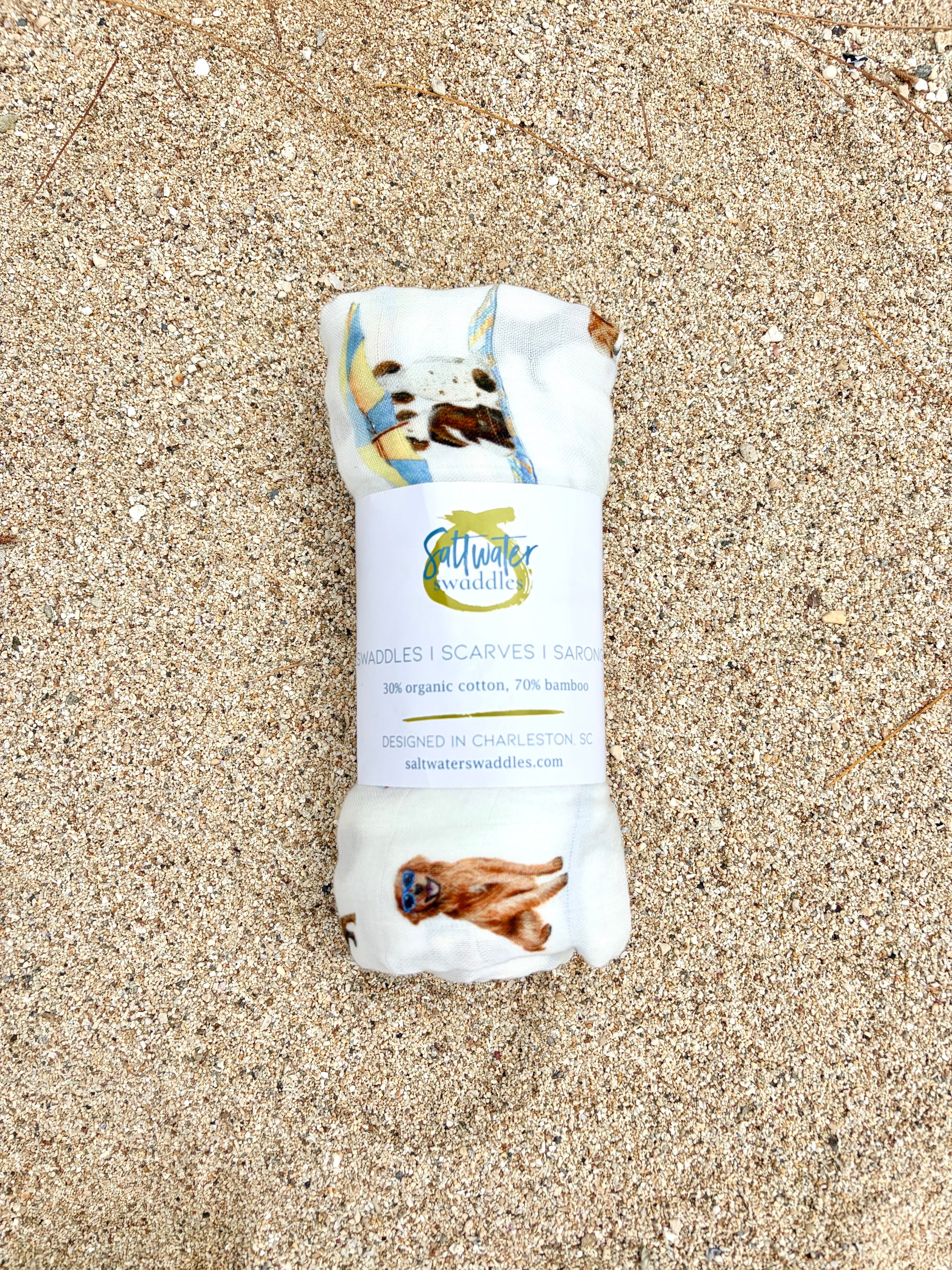 Dog Days of Summer pattern swaddle displayed rolled with the Saltwater Swaddles label on beach sand.