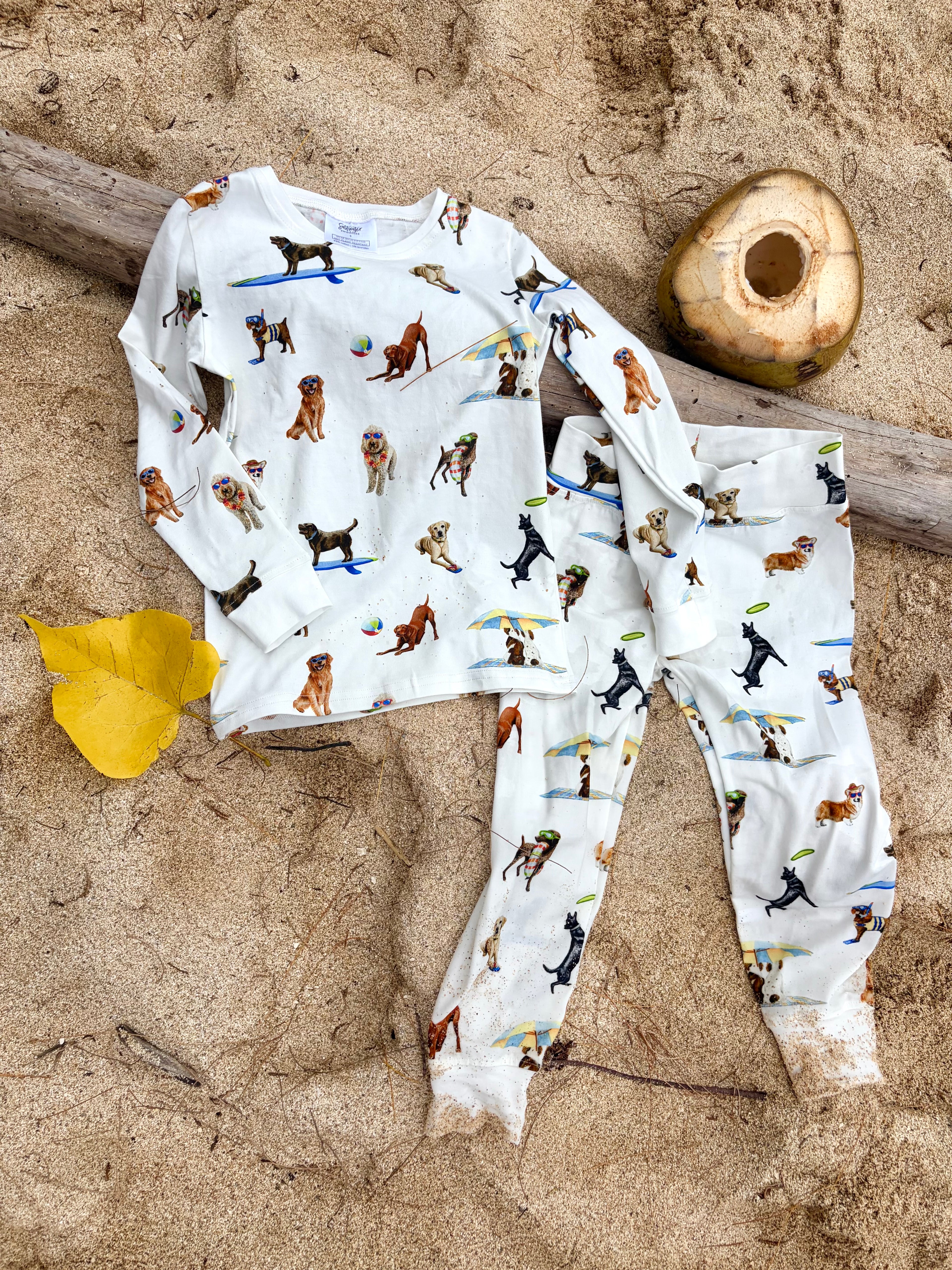 Dog Days of Summer Kids Pajamas, a snug-fitting two-piece pajama set includes a long-sleeve top and matching pants with a wide, comfortable waistband, stretchy fabric, and fitted wrist and ankle cuffs. Displayed flat on beach sand outdoors.
