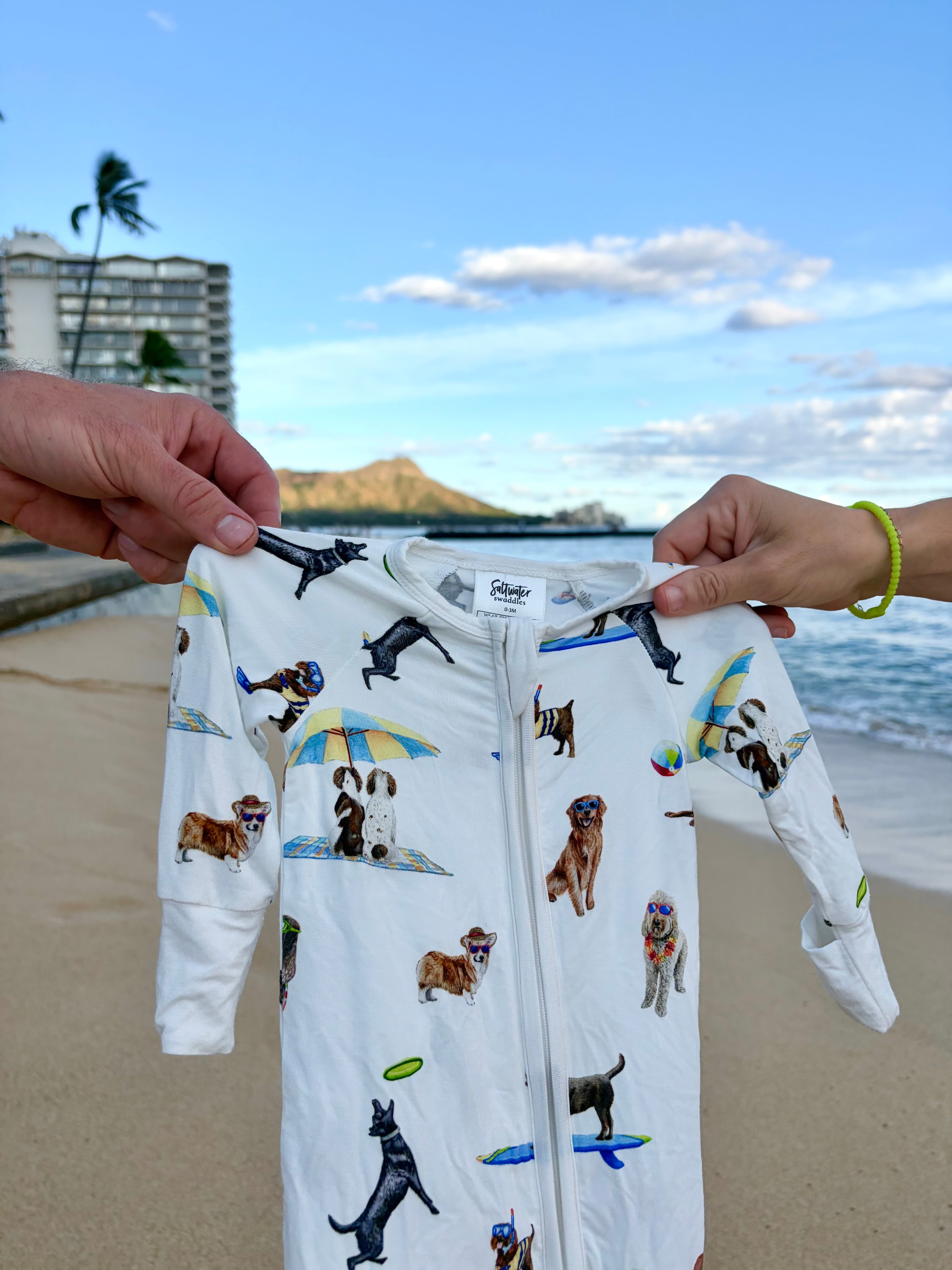 Captured against a scenic, sunny coastal backdrop featuring a sandy beach, ocean, boardwalk, and a prominent mountain, the white zippered baby romper is held up by two hands sporting yellow and green beaded bracelets. The garment itself features a playful "Dog Days of Summer" print with various dog breeds engaging in beach activities like surfing, snorkeling, and relaxing under umbrellas, all completed with comfortable white ribbed cuffs at the wrists and ankles.