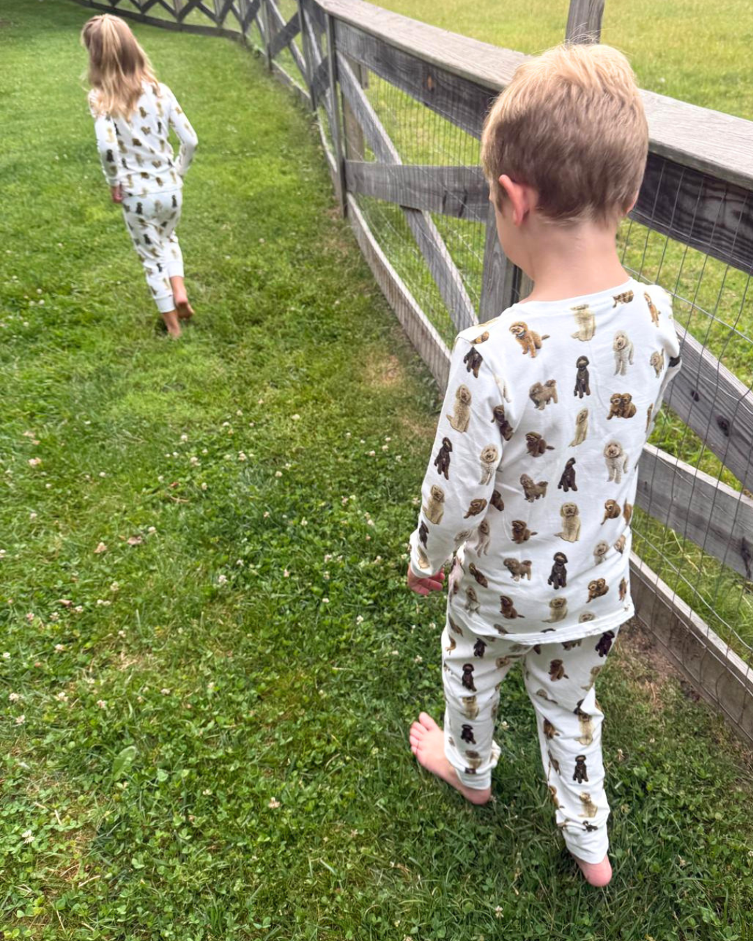 Two young children playing together outdoors, wearing matching two-piece white pajama sets patterned with hand-painted goldendoodle dogs.