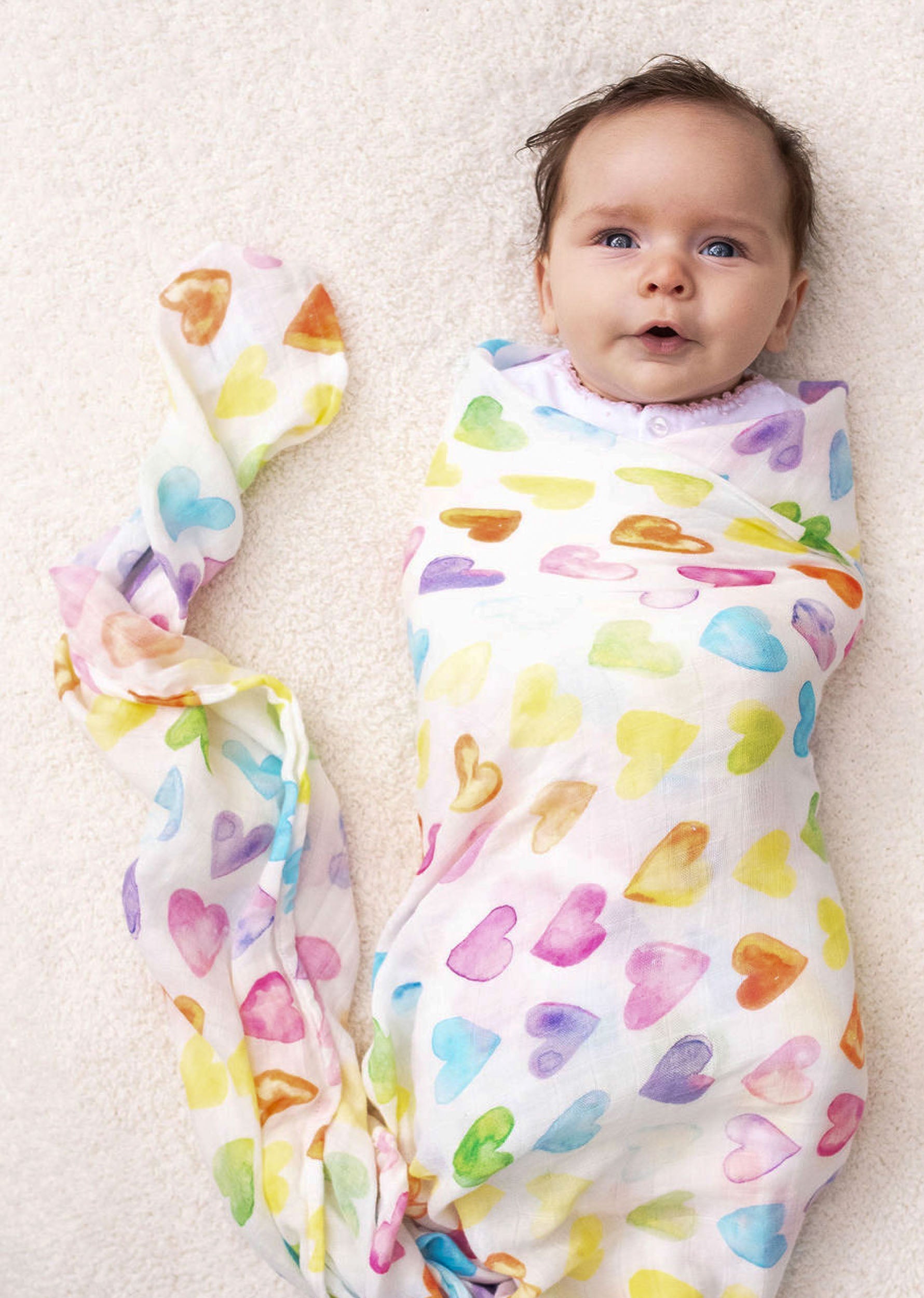 Baby wrapped in a colorful All My Love Swaddle lying on an off-white fur blanket. A soft baby blanket that has a rainbow-colored heart pattern.