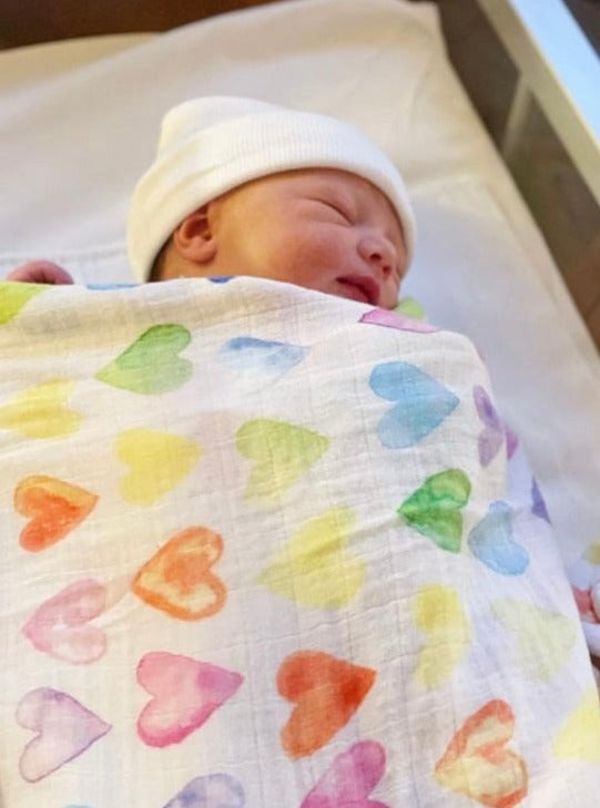 A baby in a soft Rainbow Row swaddle blanket made from organic white cotton and bamboo, adorned with rainbow-colored heart designs. 