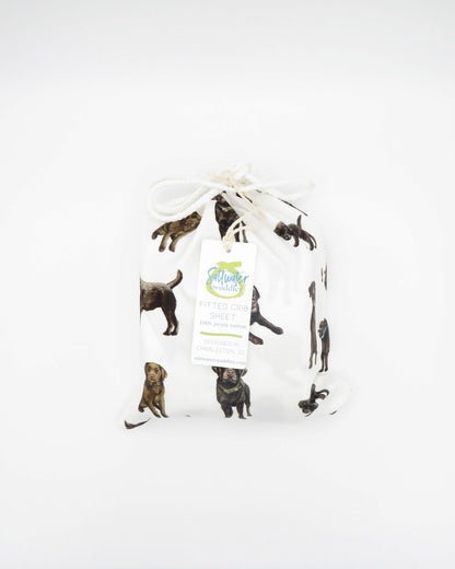 A white crib sheet with a chocolate labrador retriever print, displayed in packaging bag tied with a ribbon on a white background.