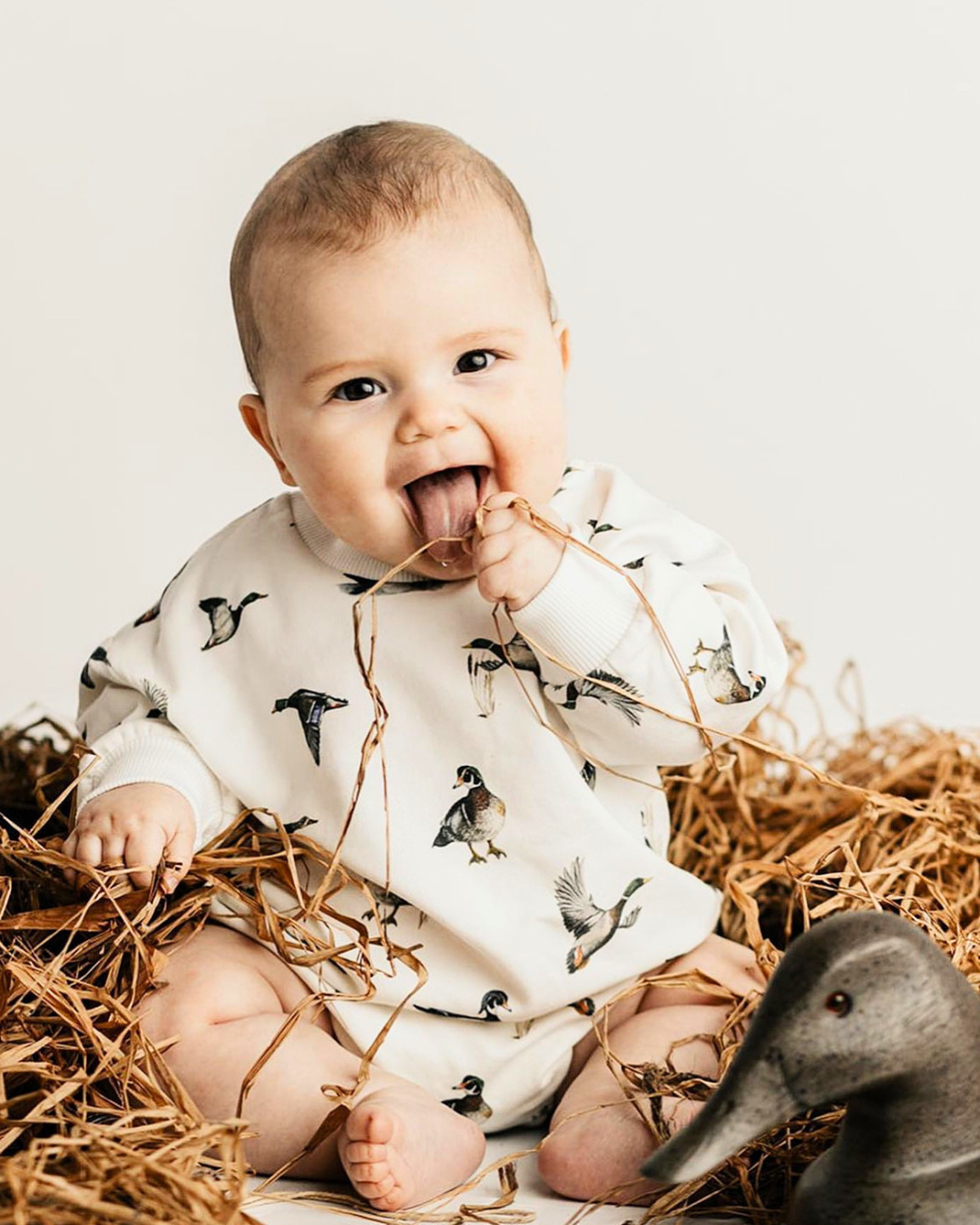 A baby wearing a long-sleeve romper with a Diving Ducks pattern, sitting in a bed of straw and posing with a wooden duck toy against a plain light background.