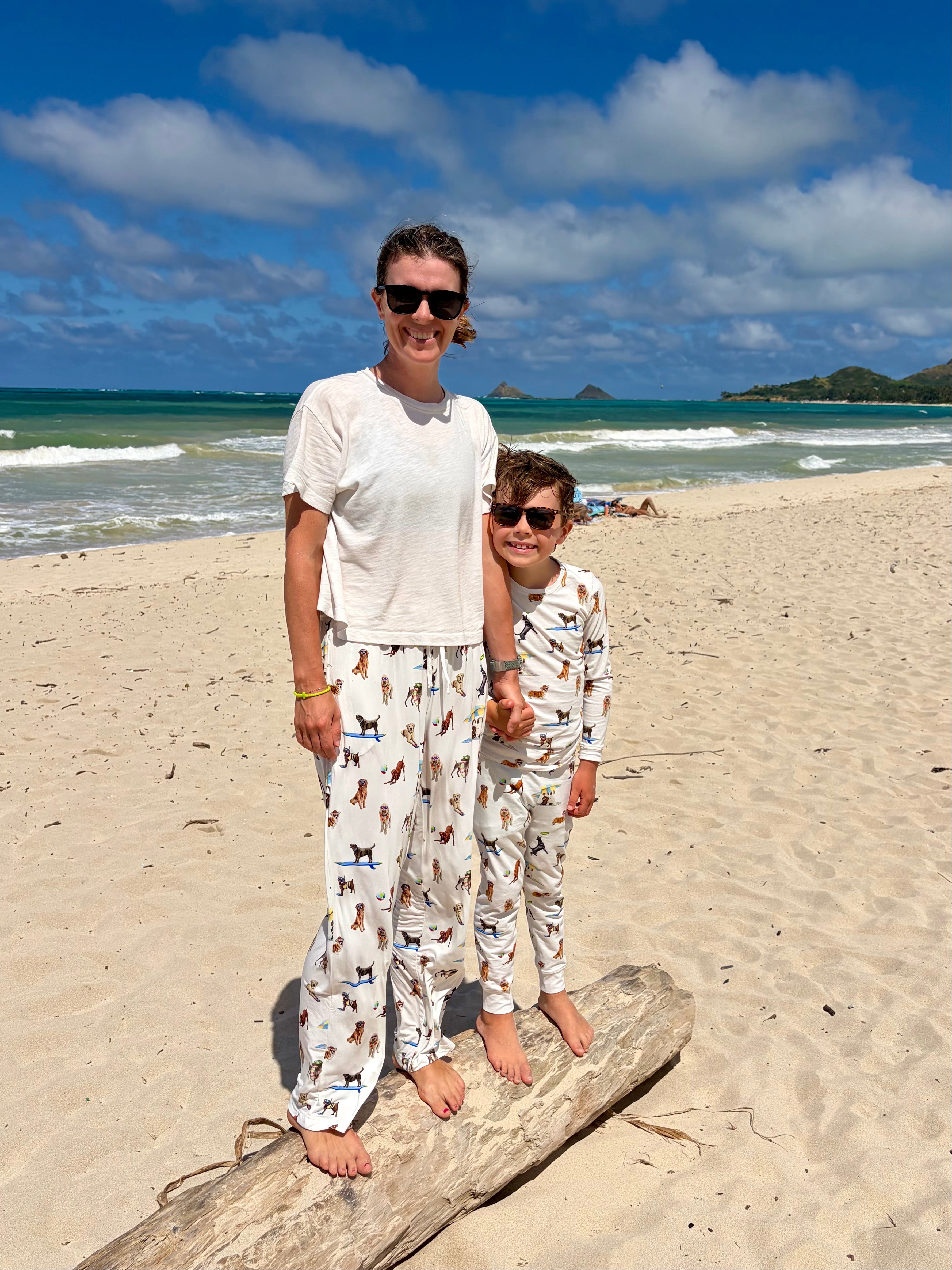 A woman and child wearing matching Dog Days of Summer pajamas stand together on a driftwood log on a sandy beach. The coordinating pajamas feature playful dog-themed illustrations. Both are wearing sunglasses, with ocean waves, blue skies, and small islands visible in the background.