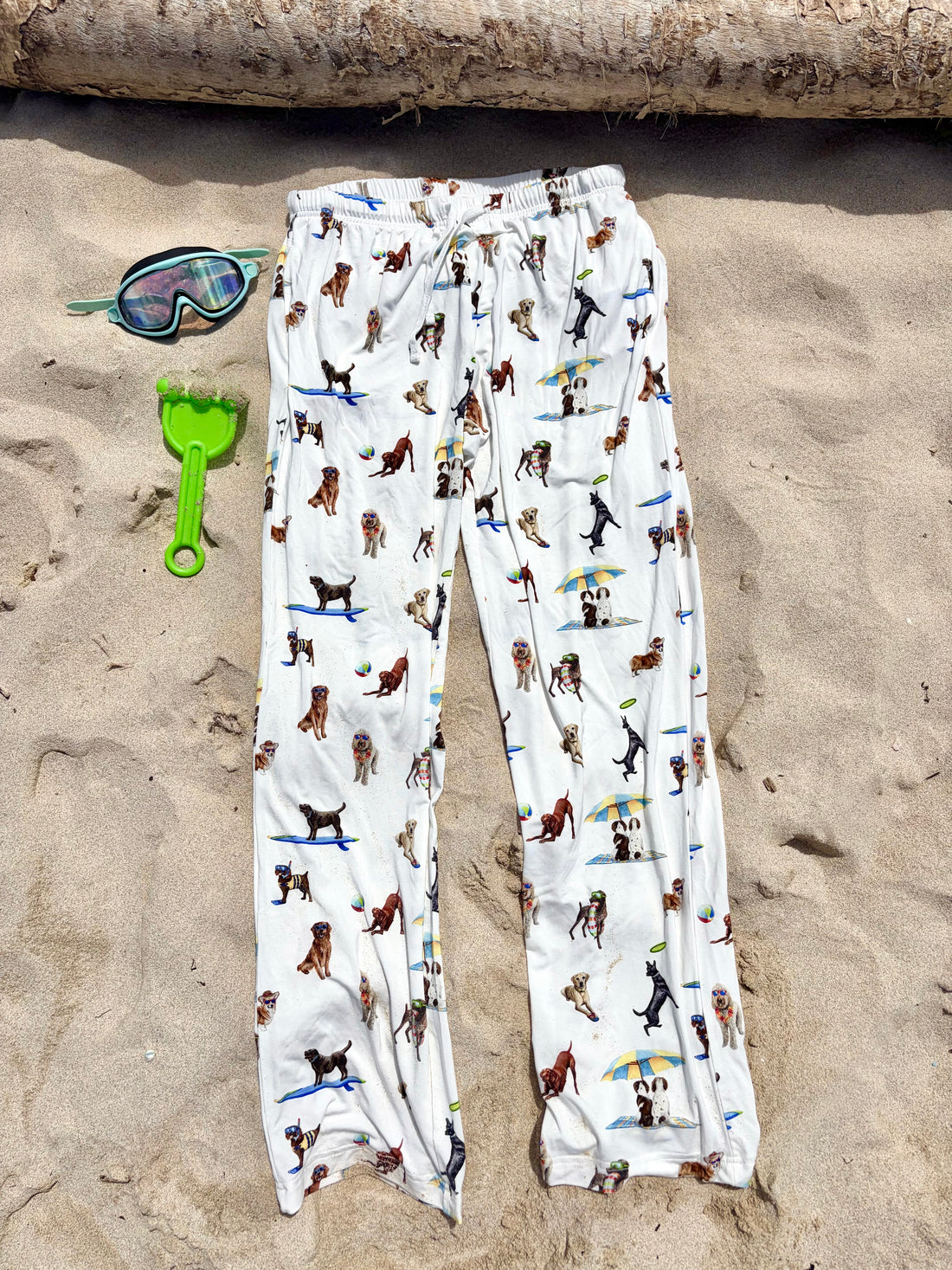 Dog Days of Summer adult pajama pants displayed flat on the beach sand, with kids’ swimming accessories on the side.
