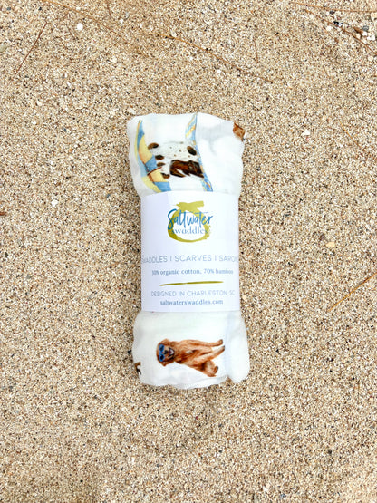 Dog Days of Summer pattern swaddle displayed rolled with the Saltwater Swaddles label on beach sand.