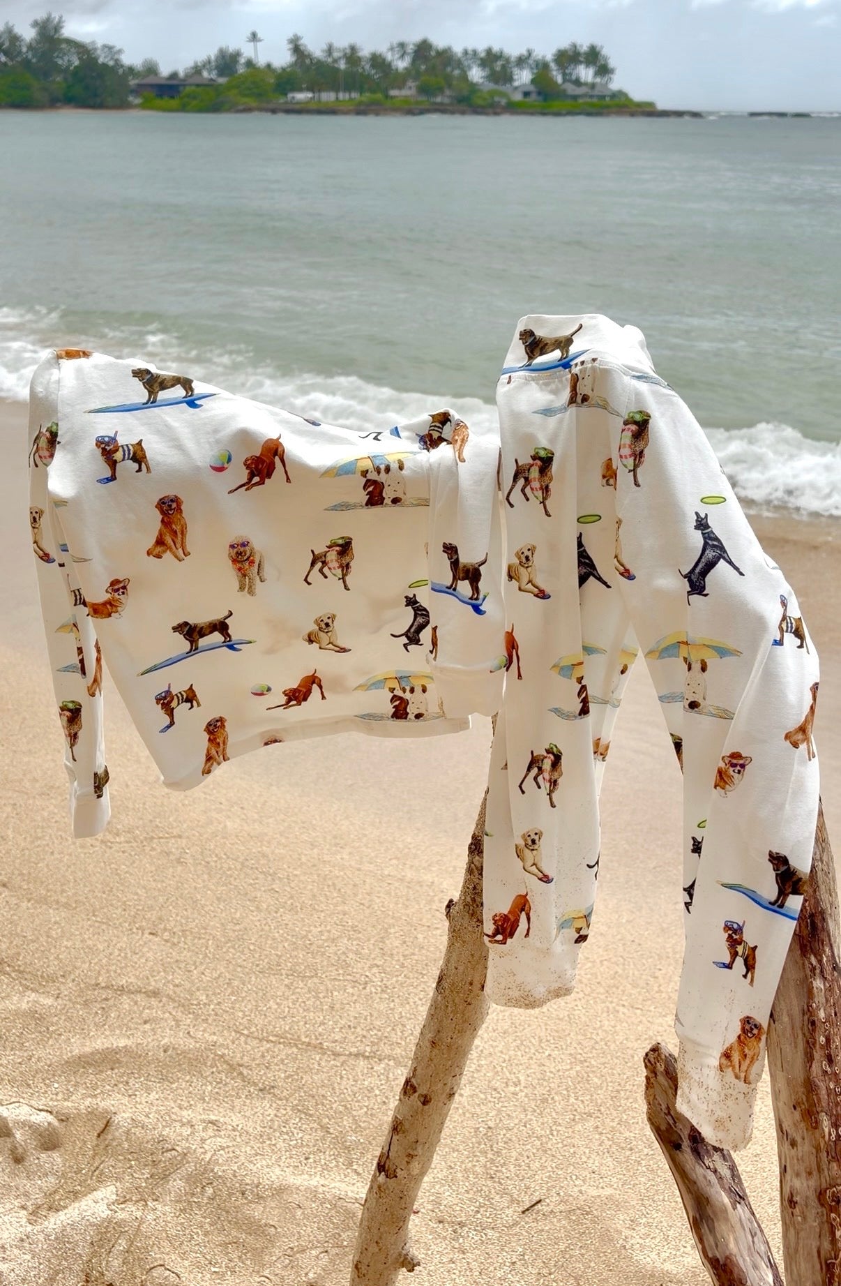 Dog Days of Summer Kids Pajamas, a snug-fitting two-piece pajama set includes a long-sleeve top and matching pants with a wide, comfortable waistband, stretchy fabric, and fitted wrist and ankle cuffs. Hung on a wooden branch by the beach