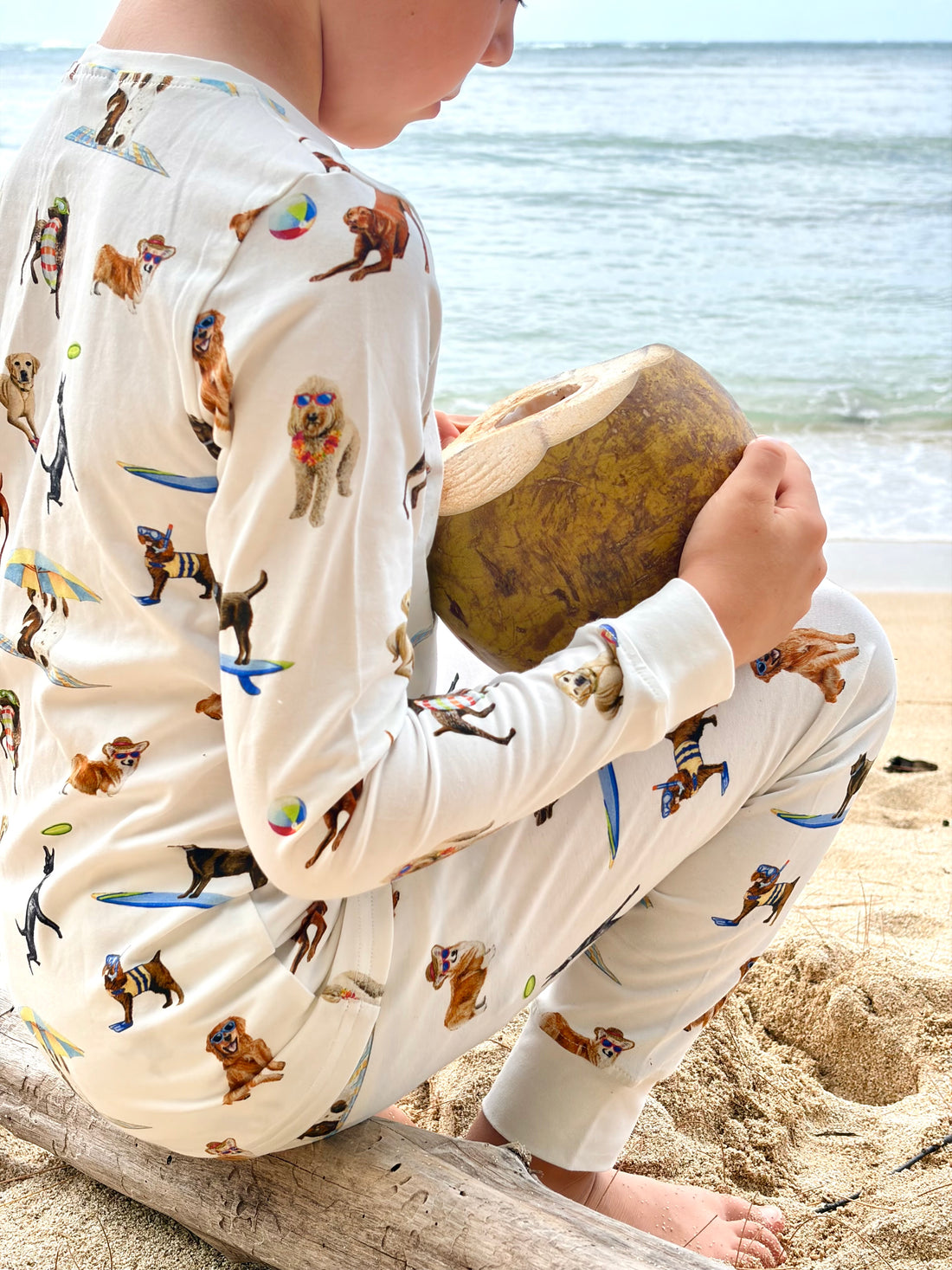 Dog Days of Summer Kids Pajamas, a snug-fitting two-piece pajama set includes a long-sleeve top and matching pants with a wide, comfortable waistband, stretchy fabric, and fitted wrist and ankle cuffs. Worn by a child at the beach while holding a coconut.