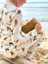 Dog Days of Summer Kids Pajamas, a snug-fitting two-piece pajama set includes a long-sleeve top and matching pants with a wide, comfortable waistband, stretchy fabric, and fitted wrist and ankle cuffs. Worn by a child at the beach while holding a coconut.