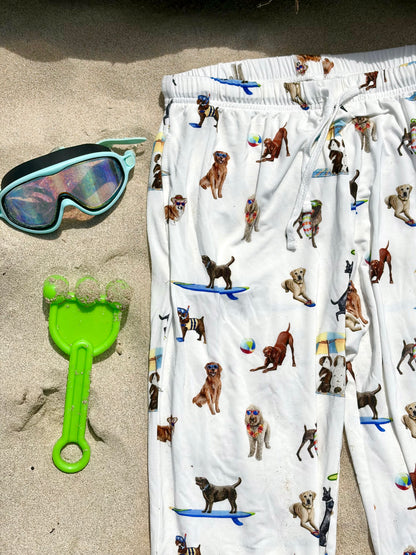 Close up look of Dog Days of Summer adult pajama pants displayed flat on the beach sand, with kids’ swimming accessories on the side.