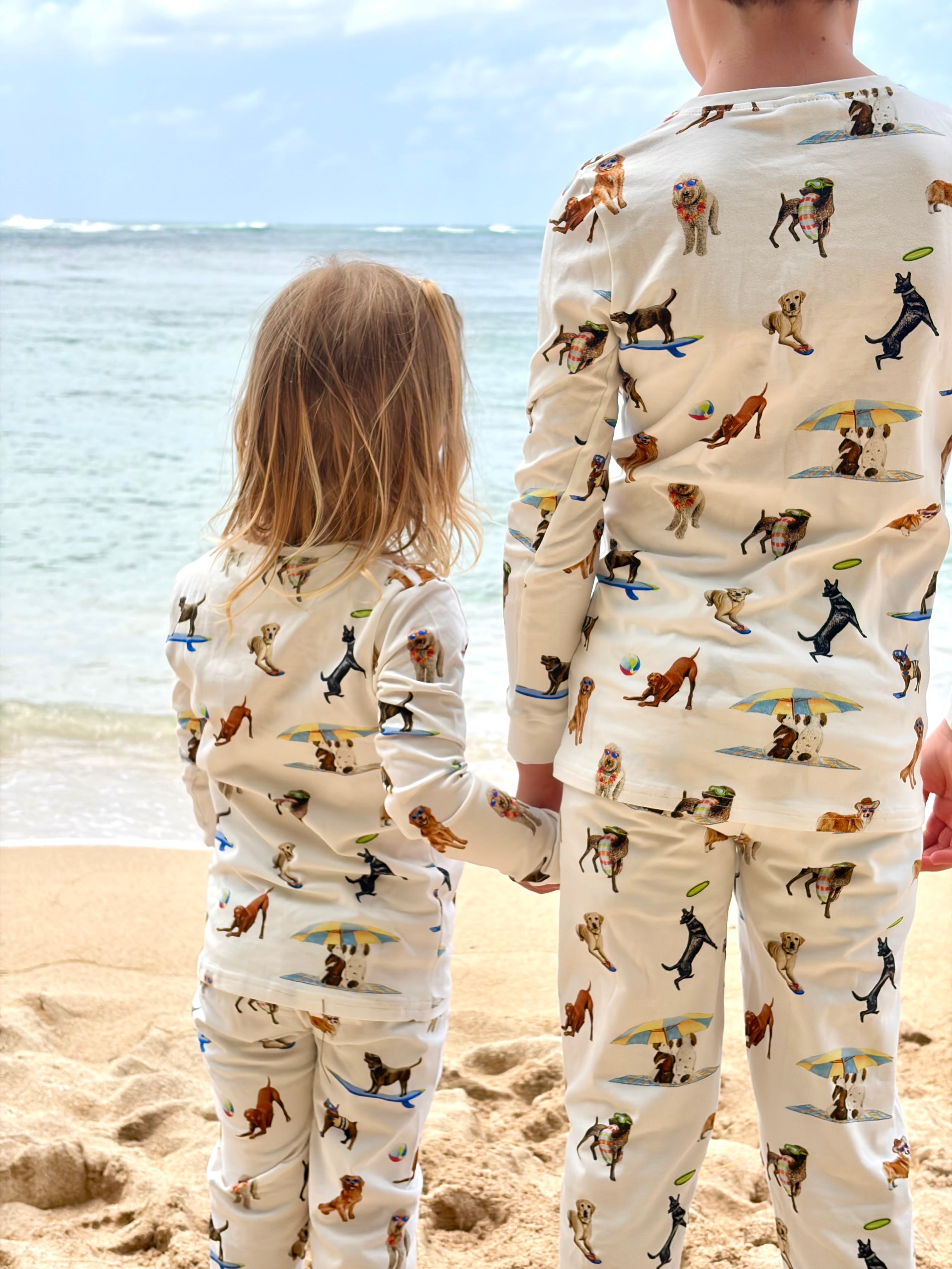 Dog Days of Summer Kids Pajamas, a snug-fitting two-piece pajama set includes a long-sleeve top and matching pants with a wide, comfortable waistband, stretchy fabric, and fitted wrist and ankle cuffs. Worn by two kids standing and facing the beach.