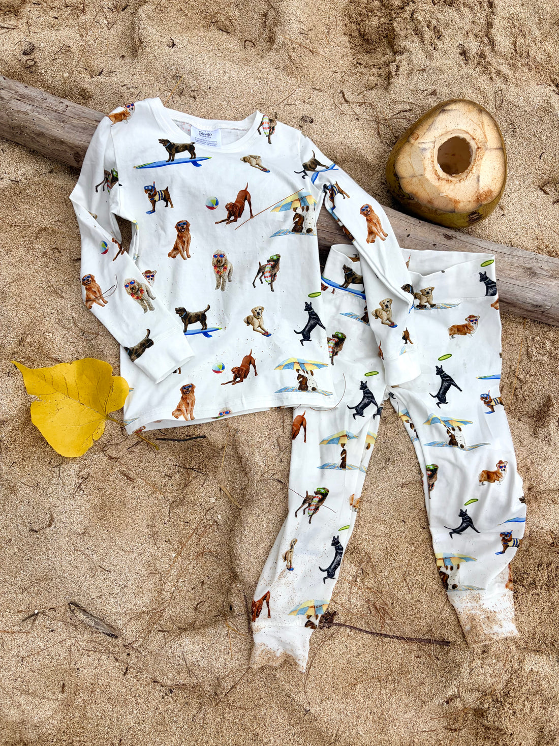 Dog Days of Summer Kids Pajamas, a snug-fitting two-piece pajama set includes a long-sleeve top and matching pants with a wide, comfortable waistband, stretchy fabric, and fitted wrist and ankle cuffs. Displayed flat on beach sand outdoors.