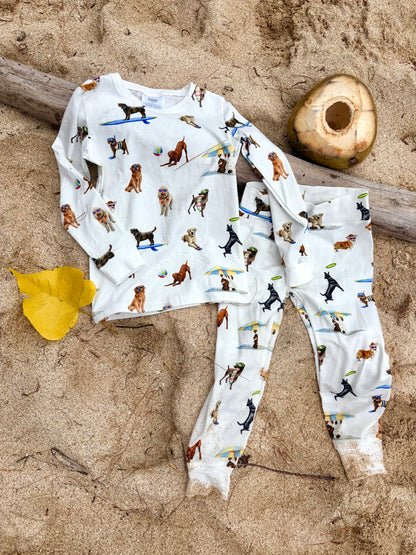 Dog Days of Summer Kids Pajamas, a snug-fitting two-piece pajama set includes a long-sleeve top and matching pants with a wide, comfortable waistband, stretchy fabric, and fitted wrist and ankle cuffs. Displayed flat on beach sand outdoors.