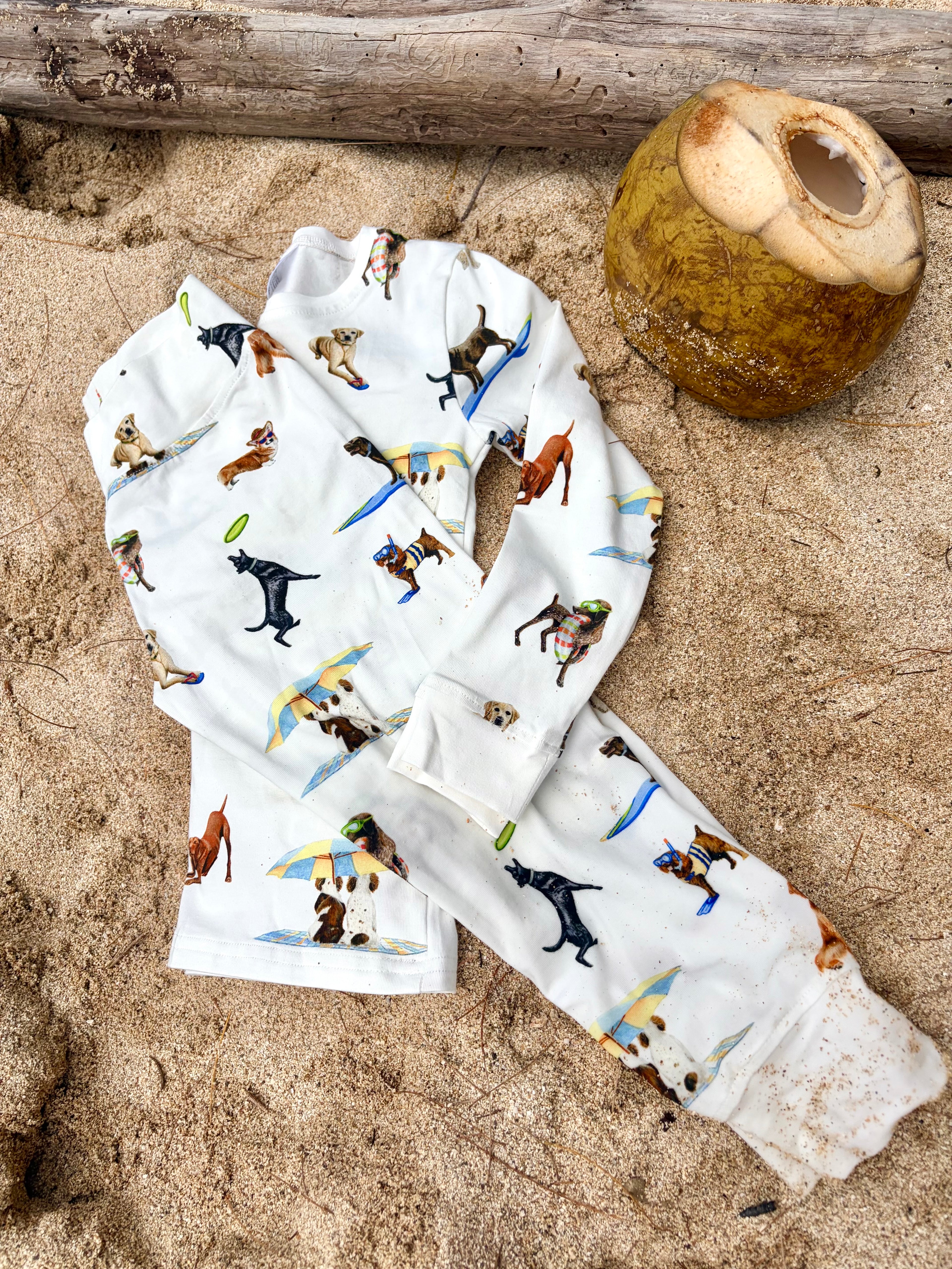 Dog Days of Summer Kids Pajamas, a snug-fitting two-piece pajama set includes a long-sleeve top and matching pants with a wide, comfortable waistband, stretchy fabric, and fitted wrist and ankle cuffs. Folded and displayed flat on beach sand outdoors.
