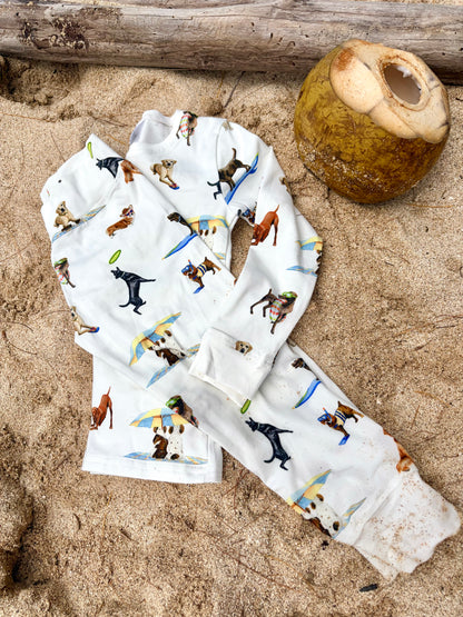 Dog Days of Summer Kids Pajamas, a snug-fitting two-piece pajama set includes a long-sleeve top and matching pants with a wide, comfortable waistband, stretchy fabric, and fitted wrist and ankle cuffs. Folded and displayed flat on beach sand outdoors.