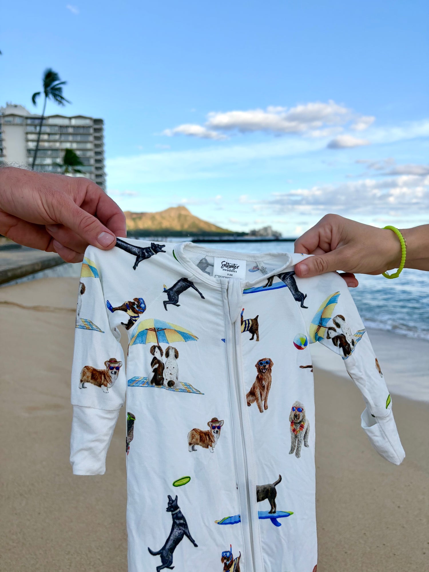 Captured against a scenic, sunny coastal backdrop featuring a sandy beach, ocean, boardwalk, and a prominent mountain, the white zippered baby romper is held up by two hands sporting yellow and green beaded bracelets. The garment itself features a playful "Dog Days of Summer" print with various dog breeds engaging in beach activities like surfing, snorkeling, and relaxing under umbrellas, all completed with comfortable white ribbed cuffs at the wrists and ankles.