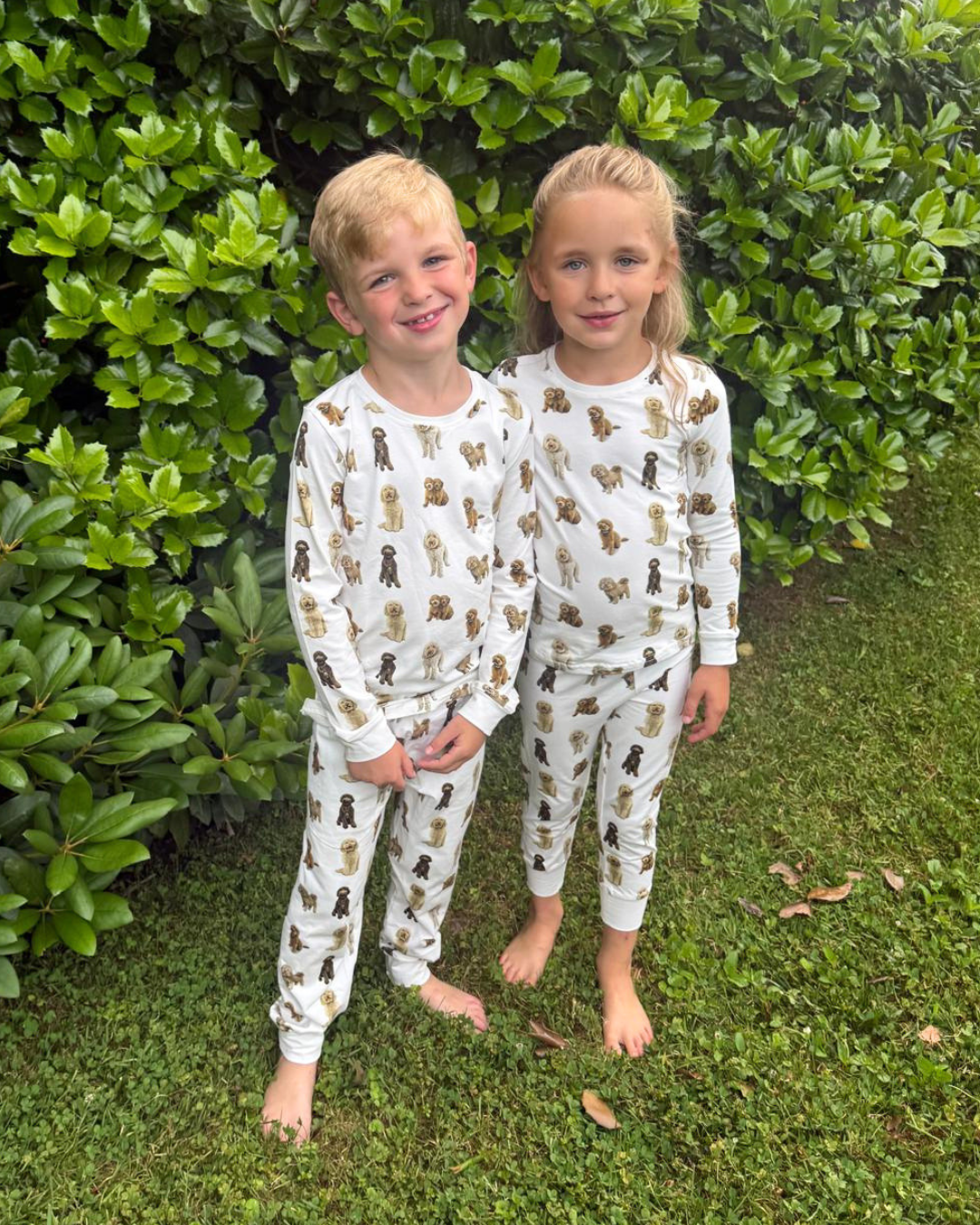 Two young children standing together outdoors smiling, wearing matching two-piece white pajama sets patterned with hand-painted goldendoodle dogs.