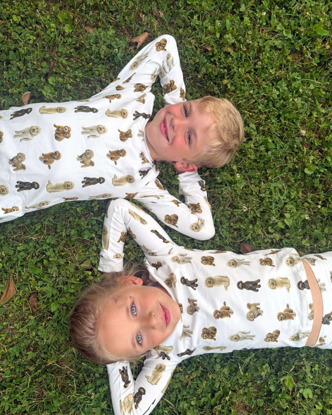 Two young children lying on the grass together outdoors smiling, wearing matching two-piece white pajama sets patterned with hand-painted goldendoodle dogs.