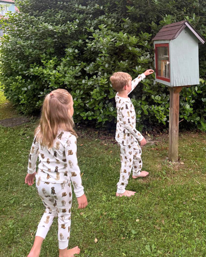 Two young children playing together outdoors, wearing matching two-piece white pajama sets patterned with hand-painted goldendoodle dogs.