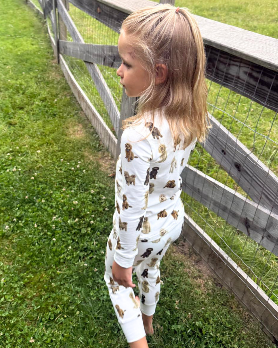A young girl standing outdoors, wearing a two-piece white pajama set patterned with hand-painted goldendoodle dogs.