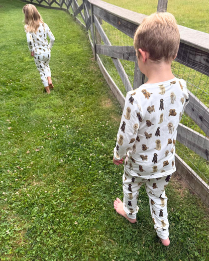 Two young children playing together outdoors, wearing matching two-piece white pajama sets patterned with hand-painted goldendoodle dogs.