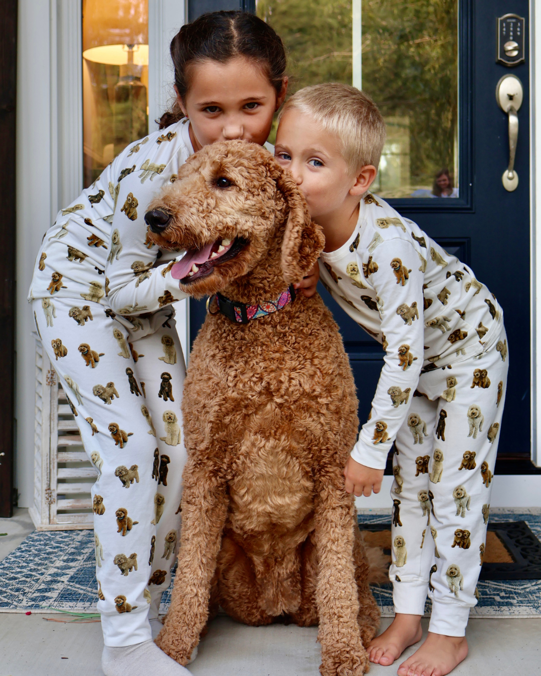 Two young children playing together outdoors, wearing matching two-piece white pajama sets patterned with hand-painted goldendoodle dogs and with their goldendoodle dog.