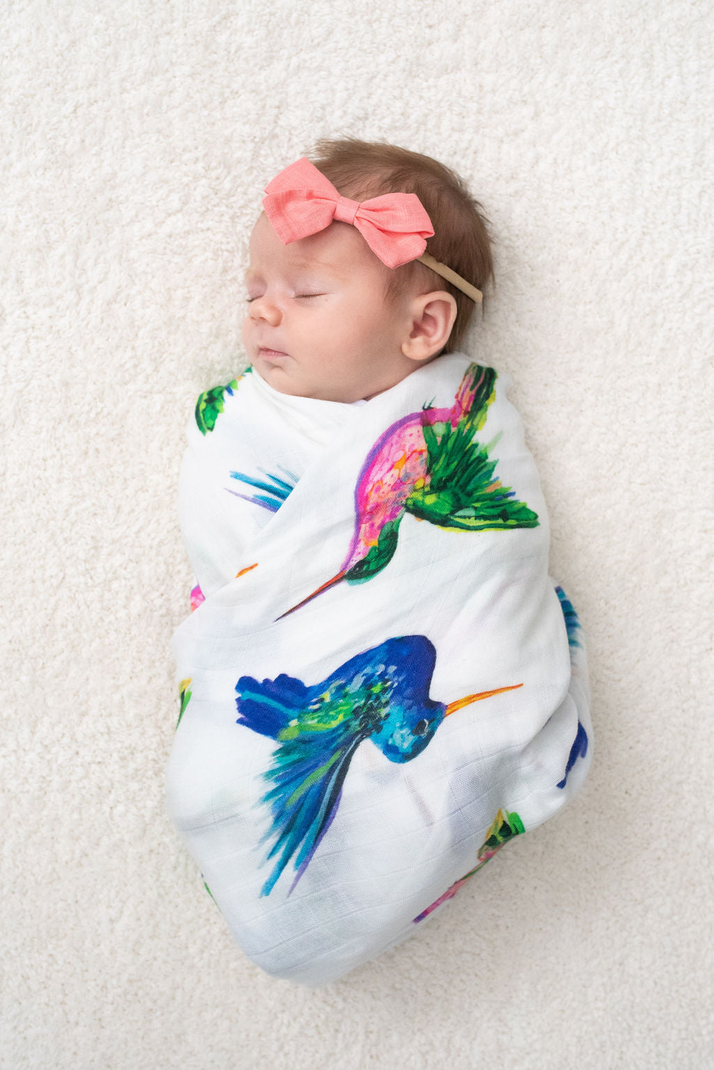 A swaddled baby with a pink headband, sleeping, and wrapped in a blanket with vibrant illustrations of hummingbirds.
