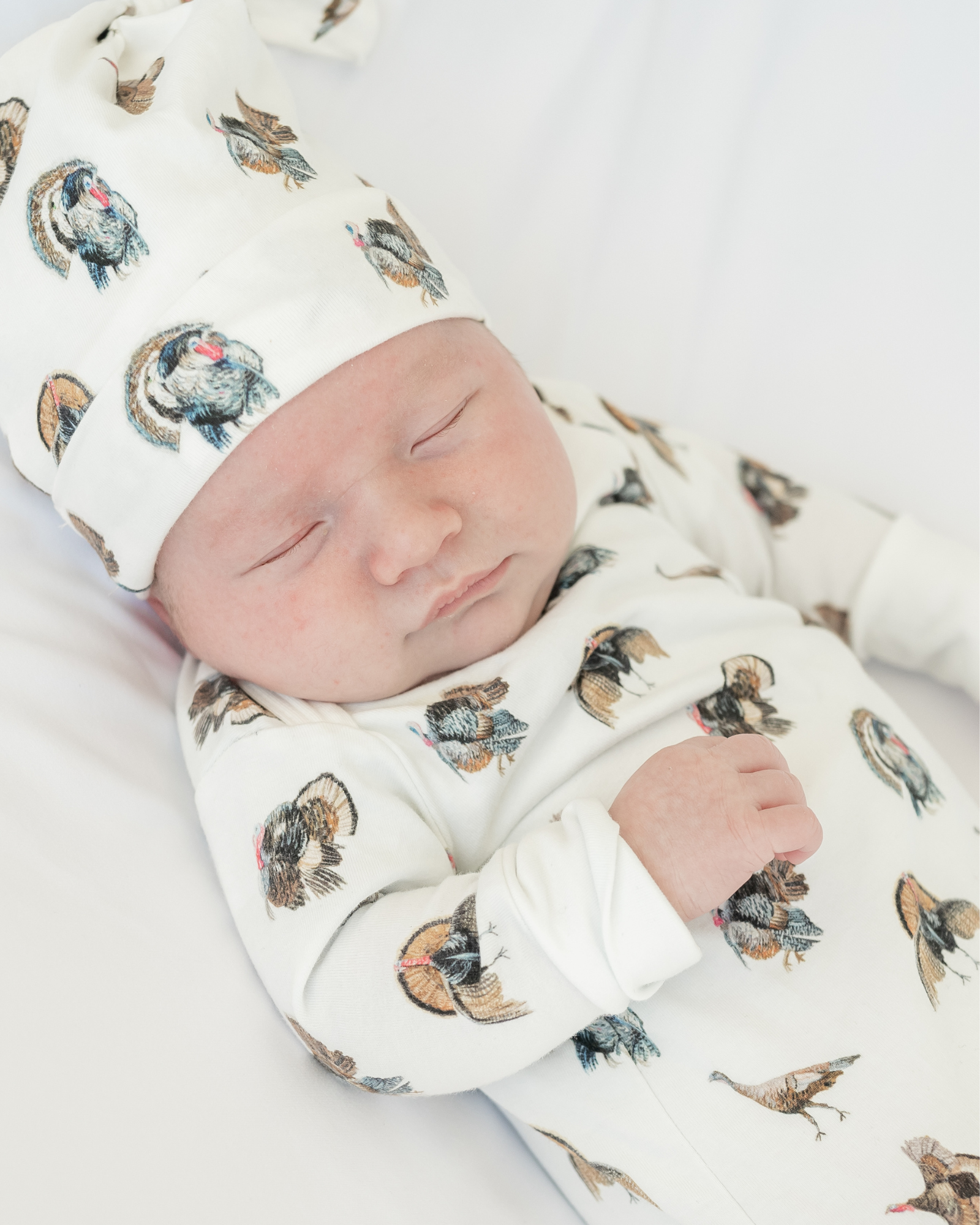 Turkey Trot newborn gown and hat set. Shown: baby peacefully sleeping in a soft, comfy gown and matching hat featuring a playful turkey design printed on soft white fabric.
