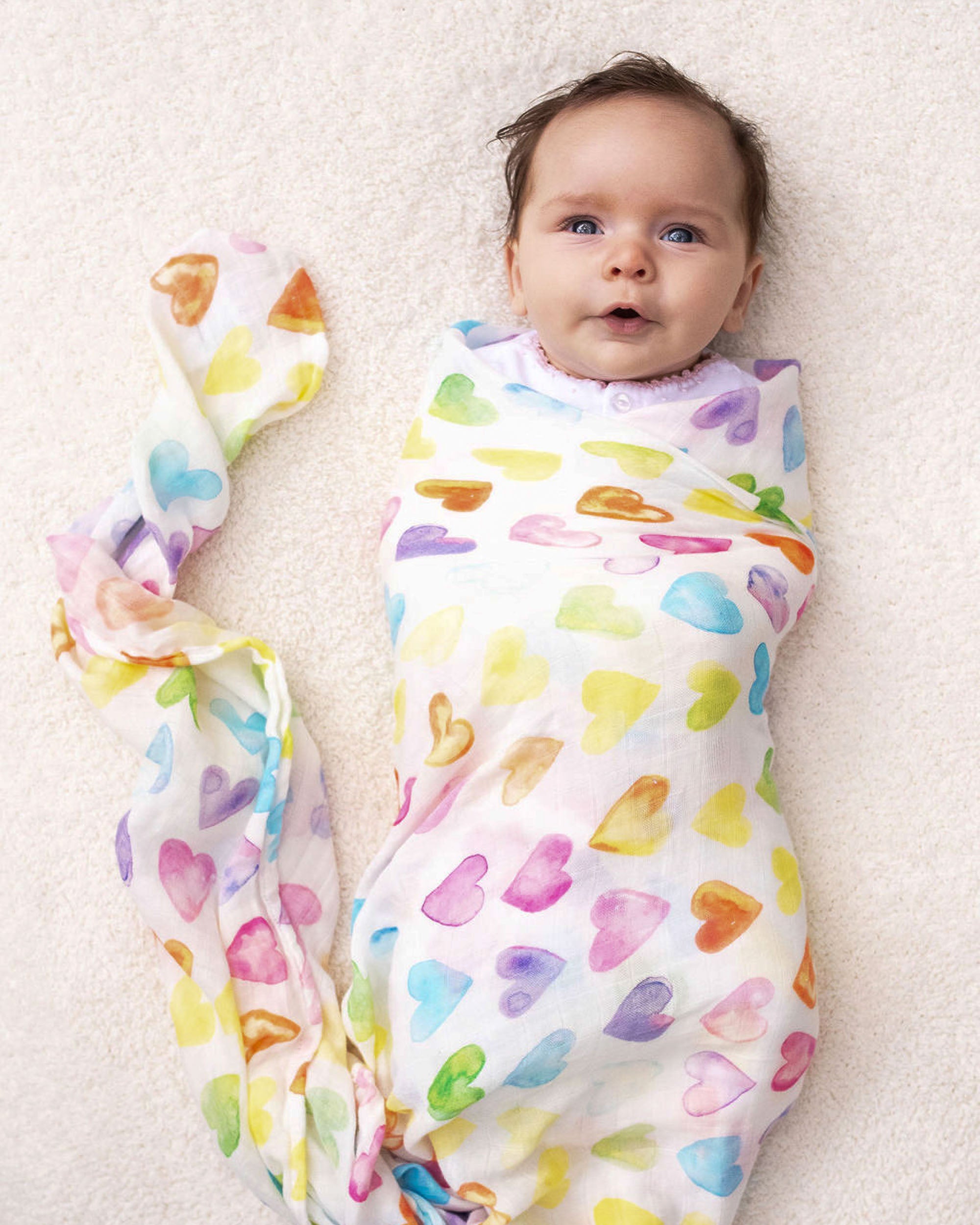 Baby wrapped in a soft Rainbow Row swaddle blanket made from organic white cotton and bamboo, adorned with rainbow-colored heart designs.