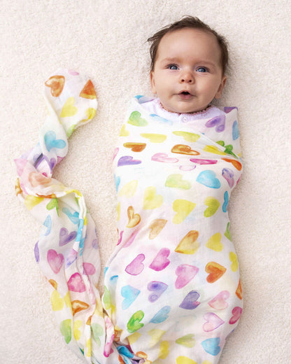 Baby wrapped in a soft Rainbow Row swaddle blanket made from organic white cotton and bamboo, adorned with rainbow-colored heart designs.