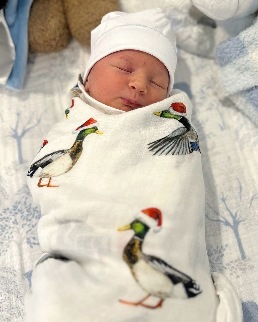 A swaddled baby with a white hat on, wrapped in a blanket with a printed design featuring christmas ducks.