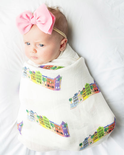 A baby wrapped in a Rainbow Row swaddle blanket, with a pink bow headband. Shown lying on a soft white fabric.