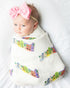 A baby wrapped in a Rainbow Row swaddle blanket, with a pink bow headband. Shown lying on a soft white fabric.