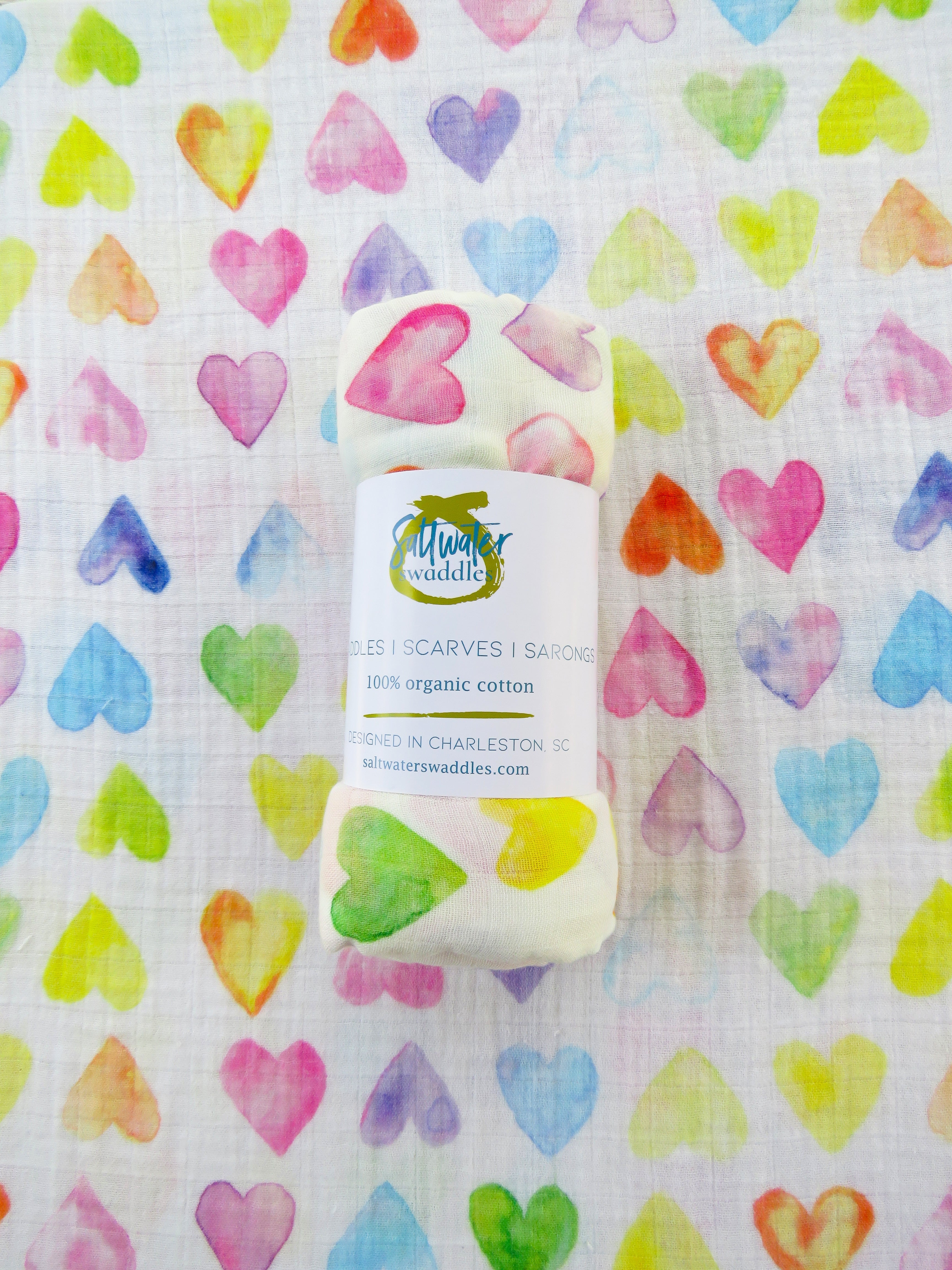 The All My Love Swaddle is displayed flat, with a rolled swaddle of the same pattern placed on top. A soft baby blanket featuring a rainbow-colored heart pattern.
