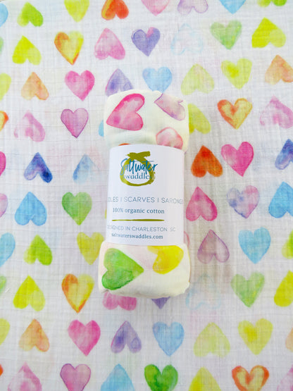 The All My Love Swaddle is displayed flat, with a rolled swaddle of the same pattern placed on top. A soft baby blanket featuring a rainbow-colored heart pattern.